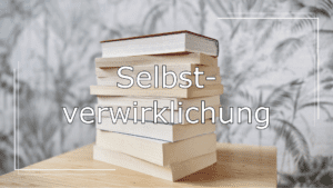 Im Hintergrund ein Stapel Bücher auf einem Holztisch. Im Vordergrund ein Schriftzug in Weiß mit schwarzer Umrandung. Der Schriftzug lautet „Selbstverwirklichung“ und verweist auf die Kategorie der Leseempfehlungen.
