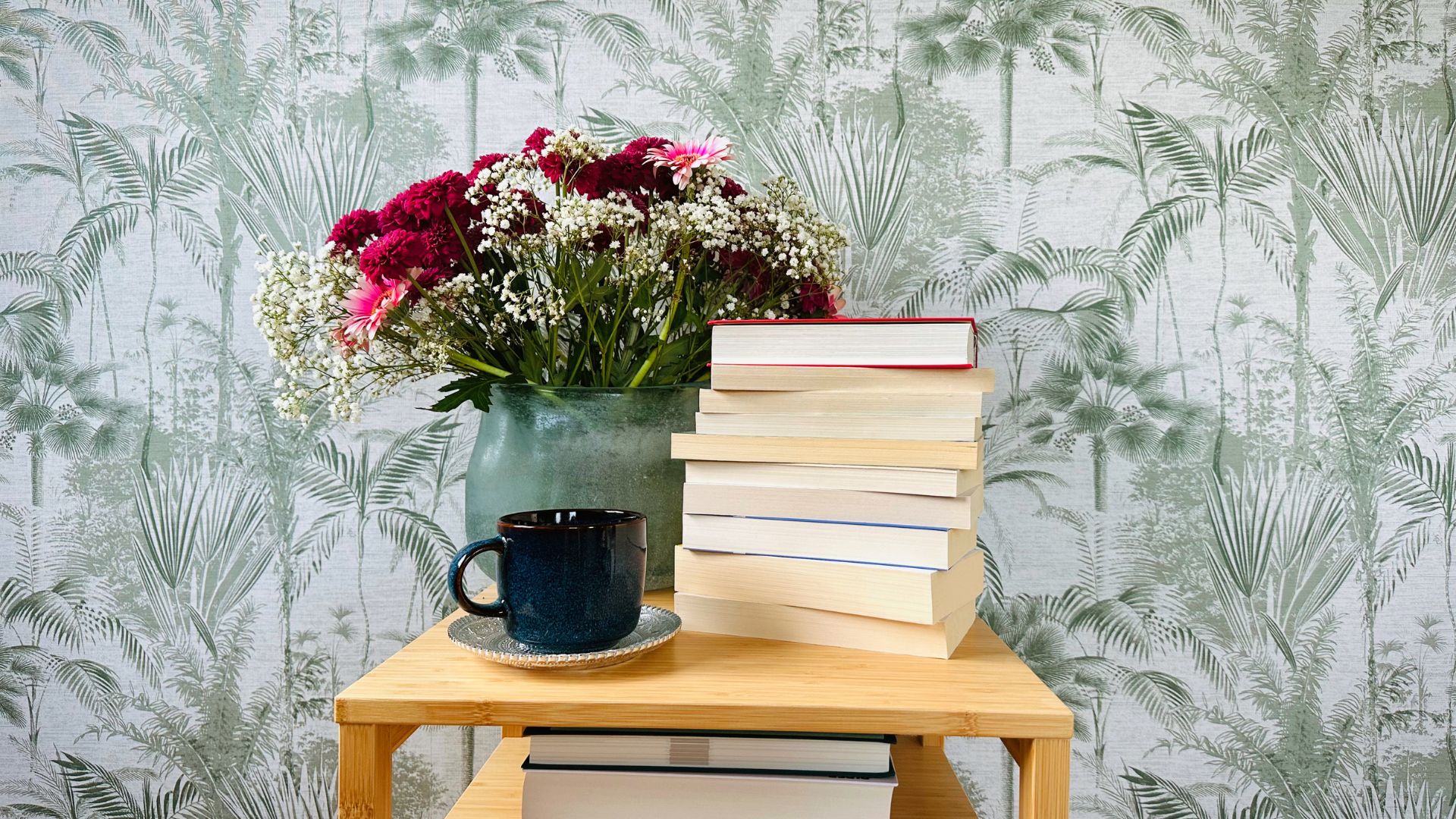 Ein gemütlicher Holztisch mit einem Stapel Bücher, einer schwarzen Tasse auf einem Untersetzer und einem Blumenstrauß in einer Vase. Im Hintergrund ist eine Tapete mit Palmenmuster zu sehen, und darüber steht das Wort 'Leseempfehlung' in geschwungener Schrift.