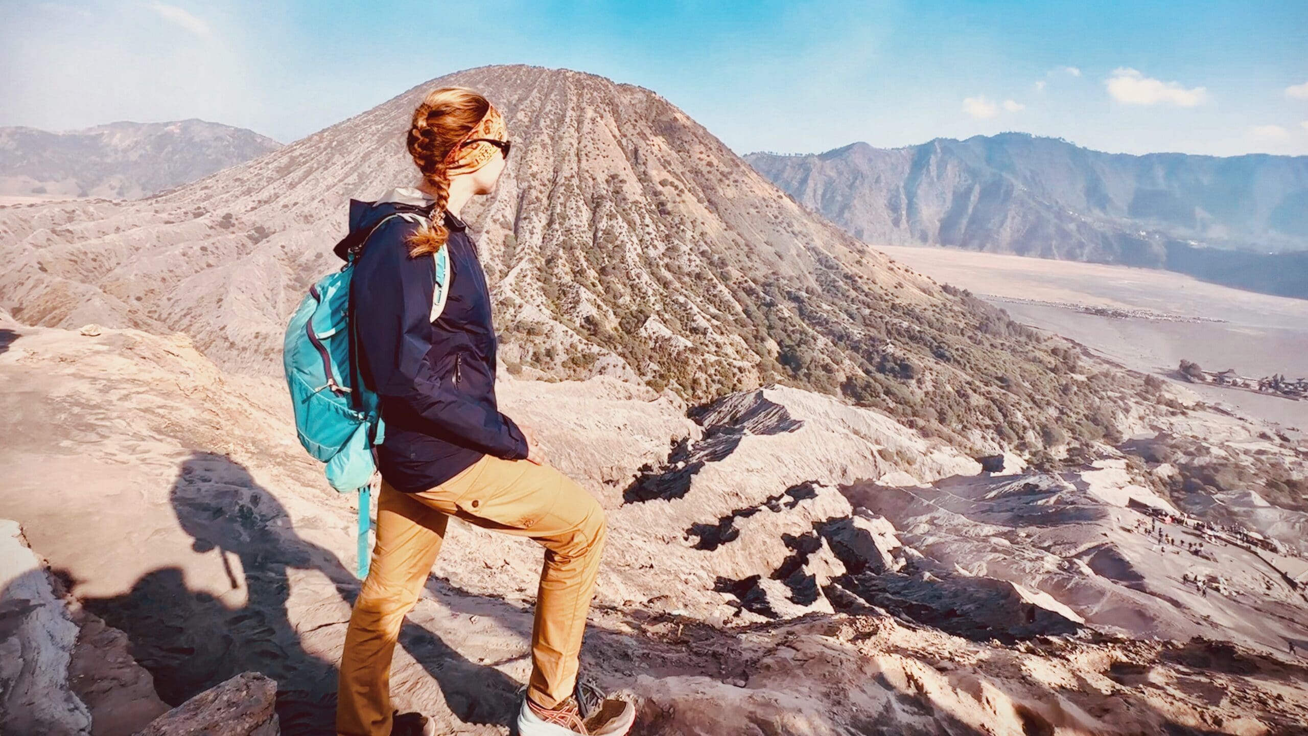 Eine Wandererin mit langem Zopf, Sonnenbrille und blauem Rucksack steht auf felsigem Untergrund mit Blick auf eine Berglandschaft. Sie hat ihre Selbstzweifel überwunden und ihr Selbstvertrauen zurückgewonnen.