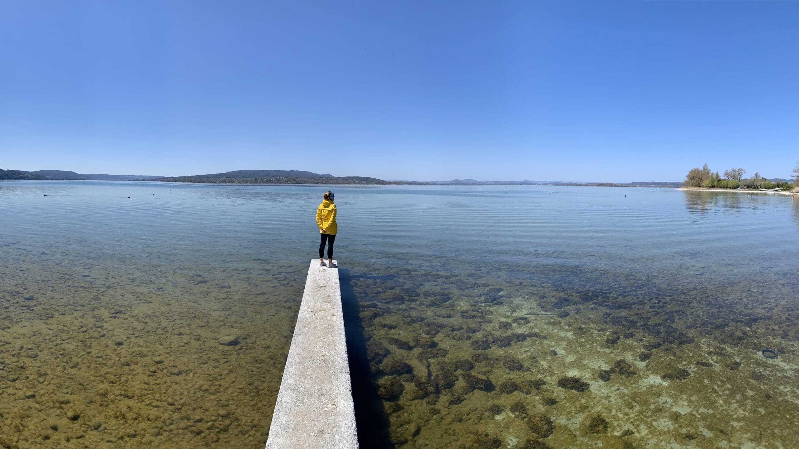 Weibliche Person in gelber Jacke steht am Ende eines Stegs und blickt auf einen ruhigen See unter klarem Himmel.