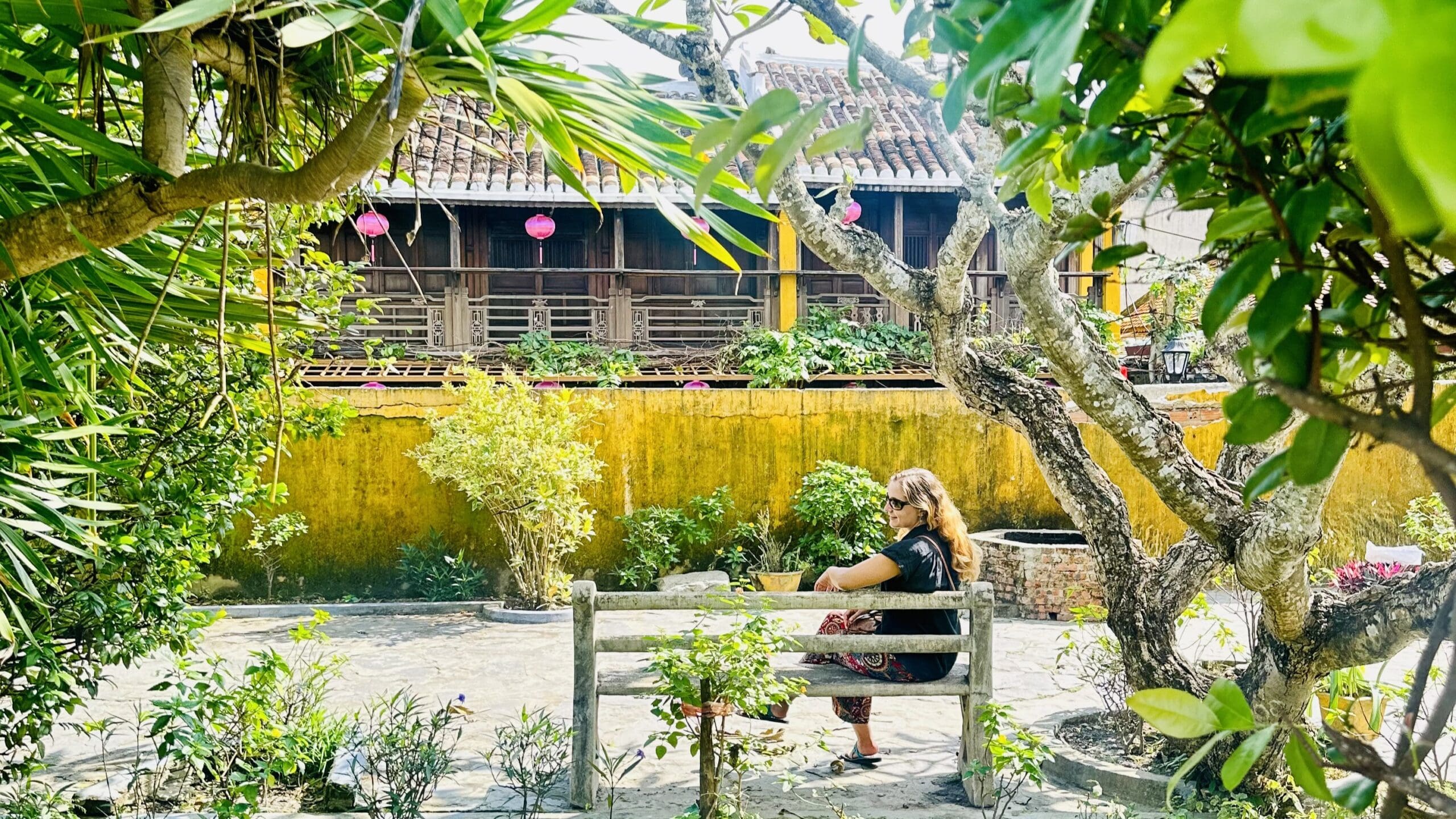 Person sitzt auf einer Bank in einem sonnigen Garten mit Bäumen und Pflanzen vor einem traditionellen vietnamesischen Haus mit gelber Wand und genießt ihr Leben ohne Social Media.