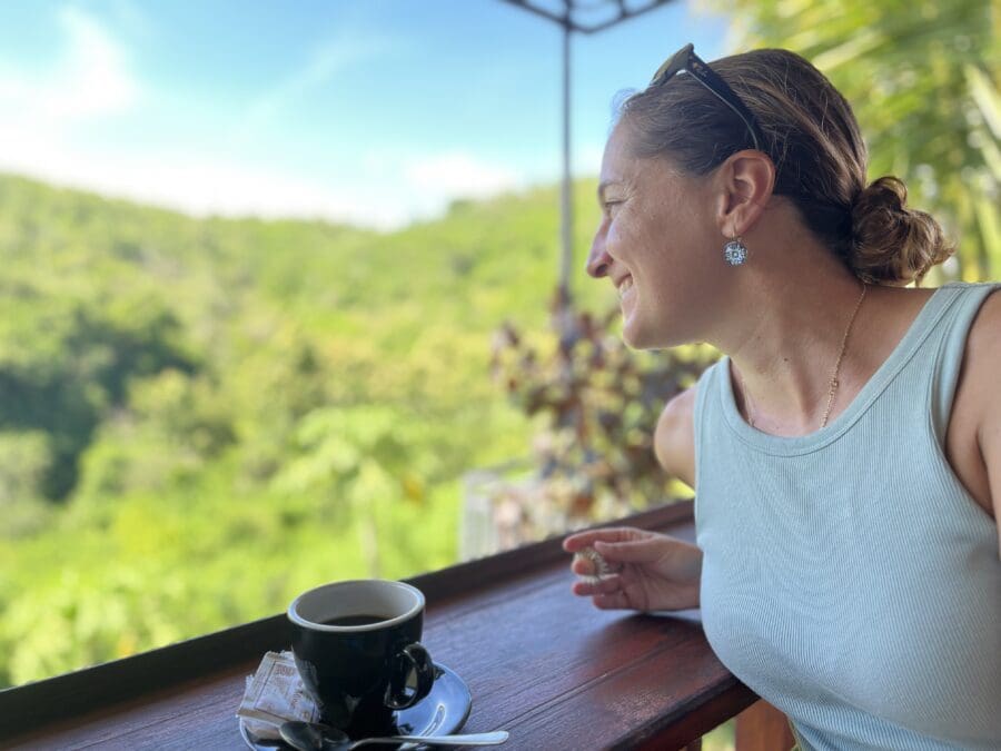 Jeanne sitzt entspannt an einem Holztisch mit Kaffee und blickt auf eine grüne Landschaft. Mit einer Social-Media-Auszeit hat man mehr Zeit für das, was wirklich zählt.